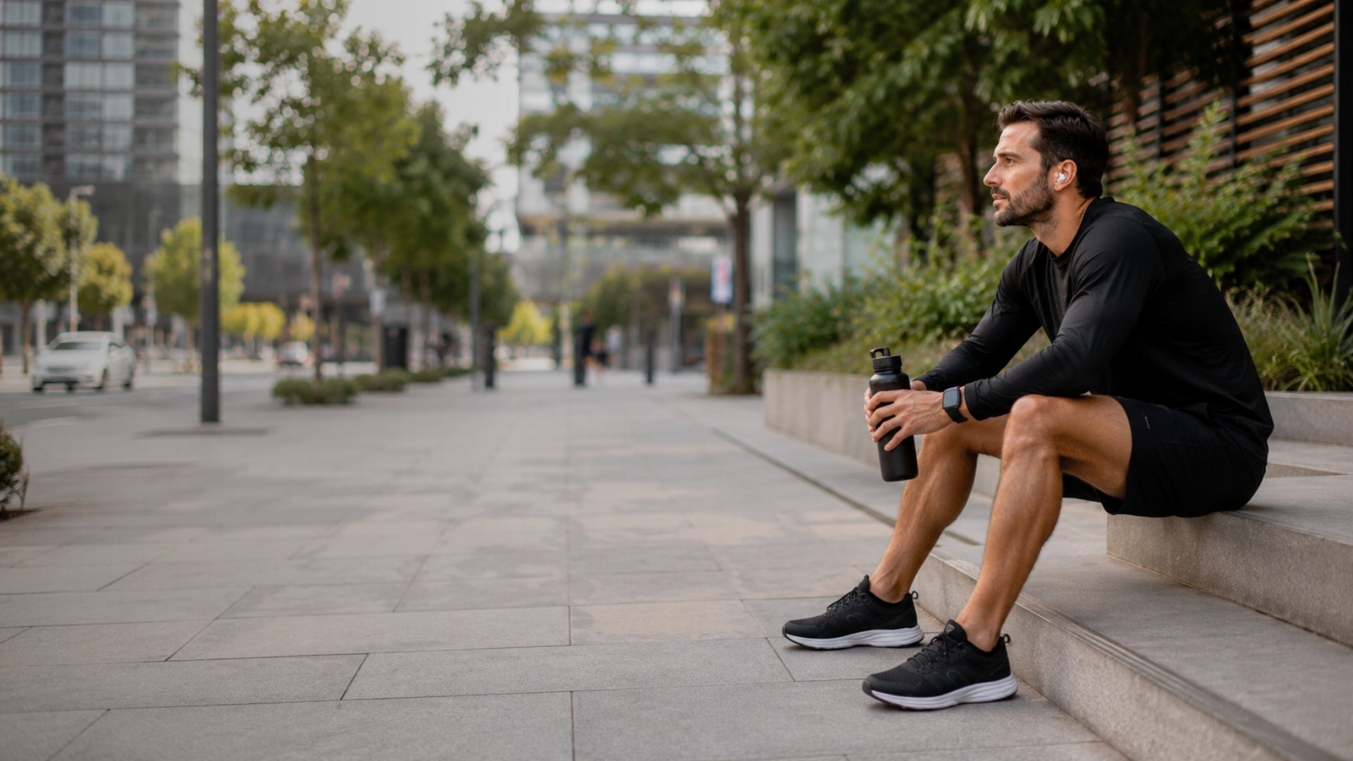 Man sitting on steps outdoors holding a water bottle, with urban setting in the background