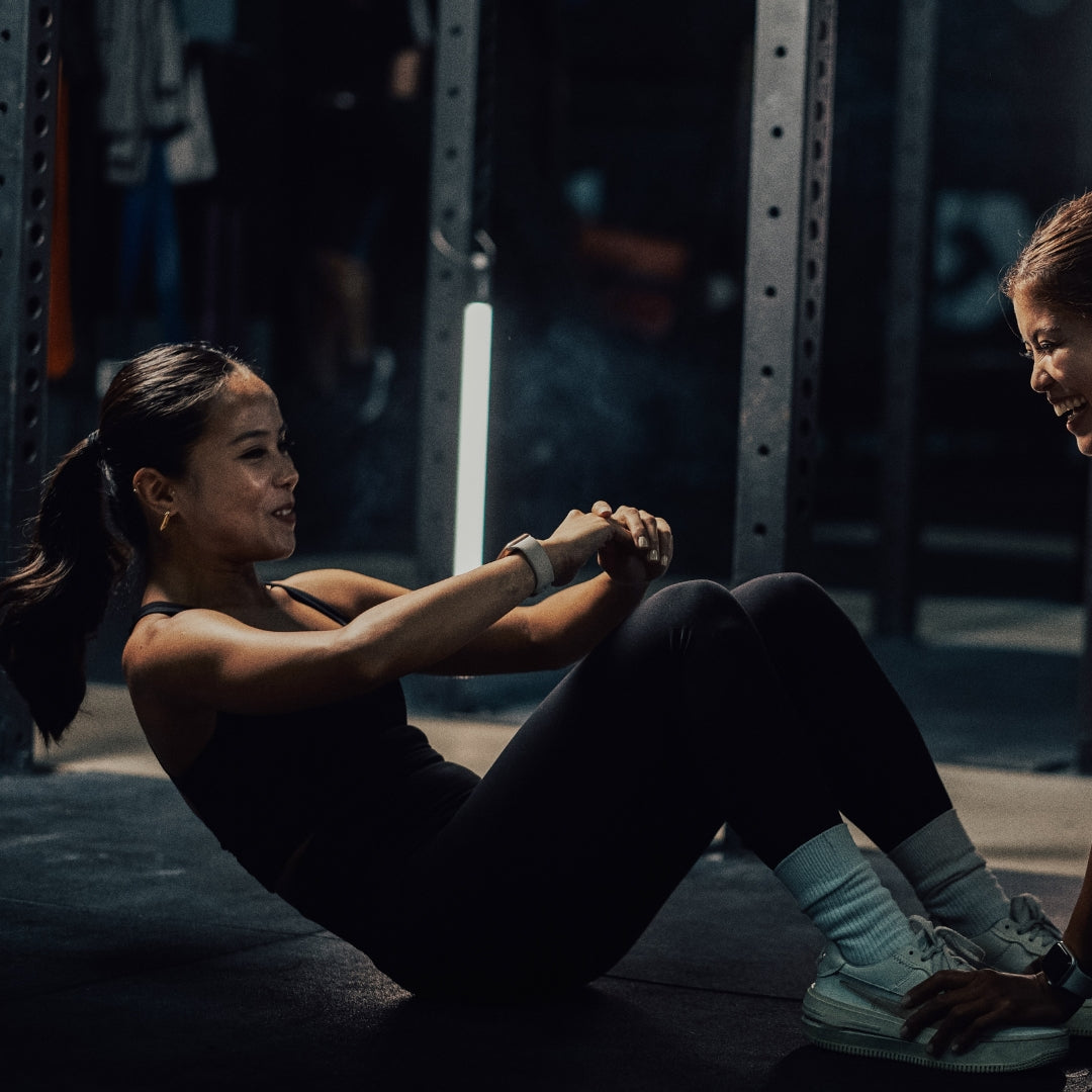 Woman exercising in a gym setting with another person partially visible.